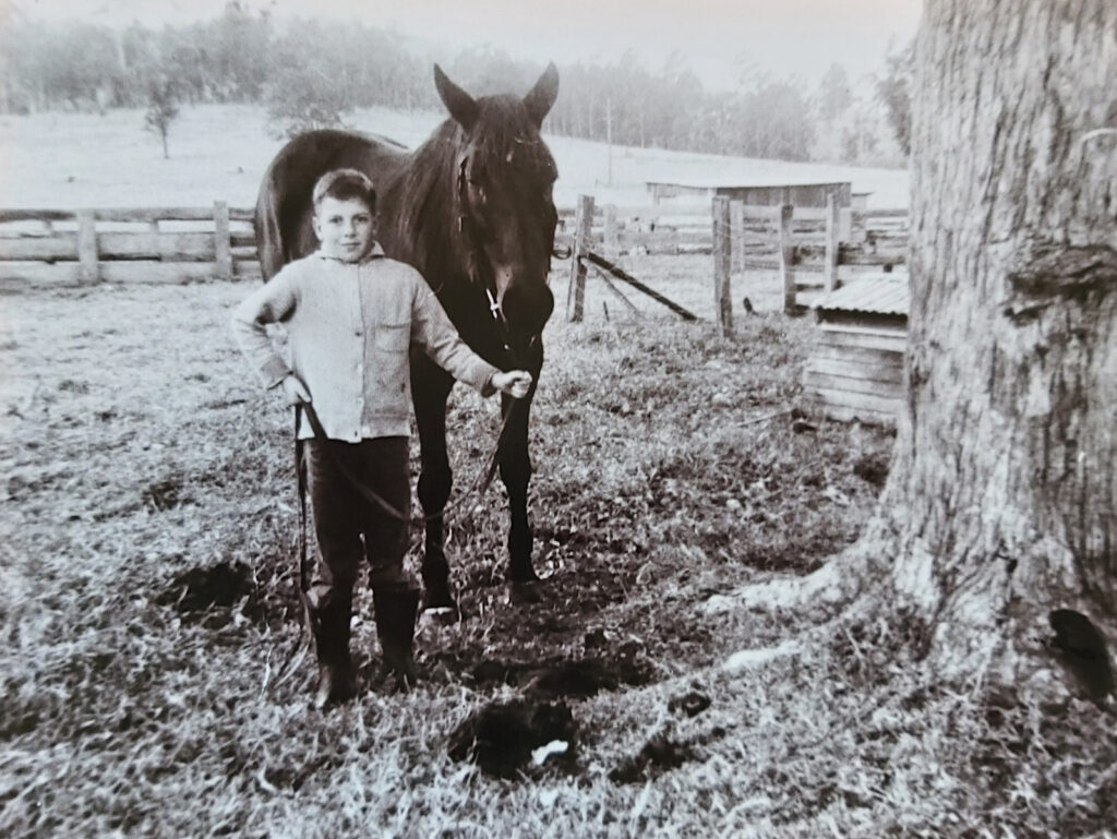 Artwork by Robbie Gambley, Victim-Survivor, when he was around 10 years old, with his bay mare, Brownie, submitted to Voices for Action. A black-and-white photograph showing a child standing beside a horse in an outdoor paddock.